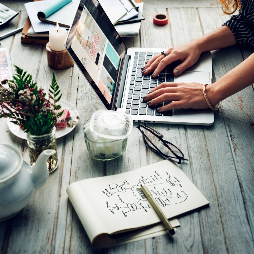 A woman focused on her laptop at a desk, surrounded by a notebook and a steaming cup of coffee.